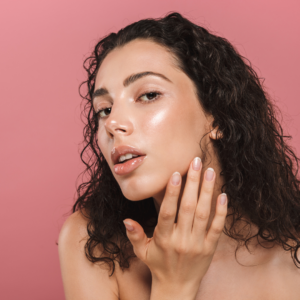 Brunette woman with clear skin posing in front of coral pink background. 