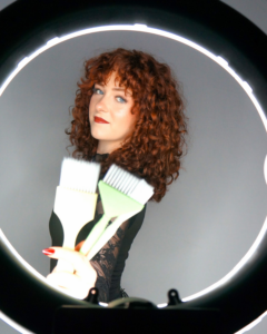 Cosmetology school student in front of ring light holding hair color brushes. 